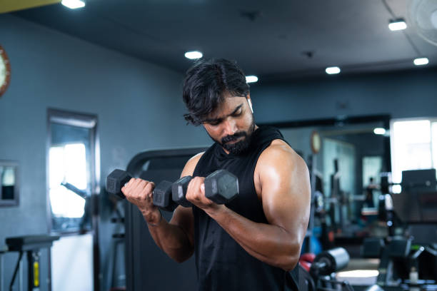 Young man exercising with dumbbells for biceps at gym - concept of bodybuilding, fitness training center, healthy lifestyle, weight lifting and recreational activity